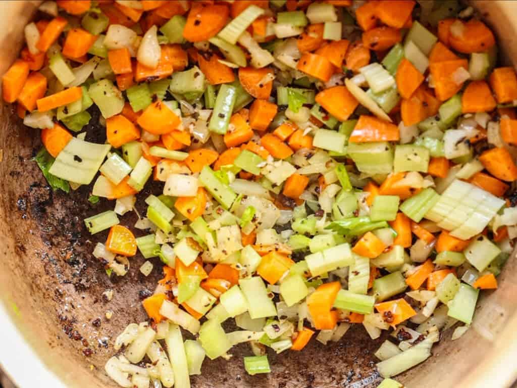 Chopped carrots, celery, and onions are sautéing in a pot, with visible seasoning and some browning on the bottom of the pan.